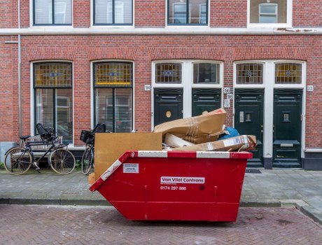 A large red skip bin filled with various household rubbish and packaging materials, positioned on a sidewalk in front of a brick terraced building with several dark green front doors and white-framed windows, some with security bars. The waste includes cardboard boxes, some flattened and others crumpled, mixed with plastic wrapping and paper waste. To the left, two bicycles are parked against the building's wall. The building has a traditional brick facade with a slightly weathered finish, and the doors and windows are evenly spaced, with some decorative yellow detailing around the window frames. The scene is set during daylight, with natural light illuminating the surface textures of the bricks, bin, and waste materials. This image relates to rubbish clearance services offered by Office Clearance Yeading in the local area, highlighting waste removal from residential properties near the postcode area associated with Yeading, UK.