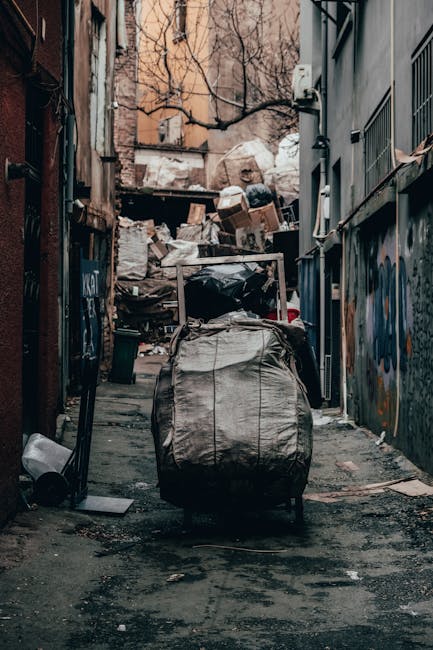 A small, old blue pickup truck parked on a quiet street with a large load of mixed debris and household rubbish piled in the truck bed and on a makeshift metal framework extending from the vehicle. Th