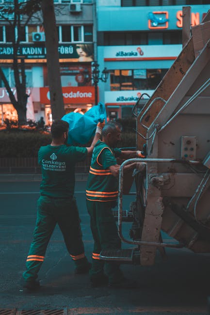 Two waste collection workers are seen operating a large, yellow skip loader vehicle on a city street during dusk or early evening. One worker, wearing an orange glove and a teal uniform with company b