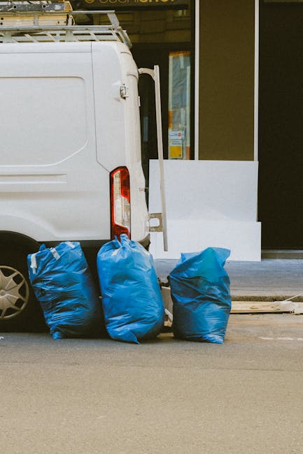The image shows the rear side of a white commercial van parked on a pavement in an urban environment. The van’s rear door is partially visible, with a vertical tail light on the left side. Positioned