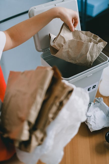 A person disposing of paper waste into a white recycling bin, with a brown paper bag in hand. The bin is placed on a wooden surface and has a partially visible label indicating it is used for waste co